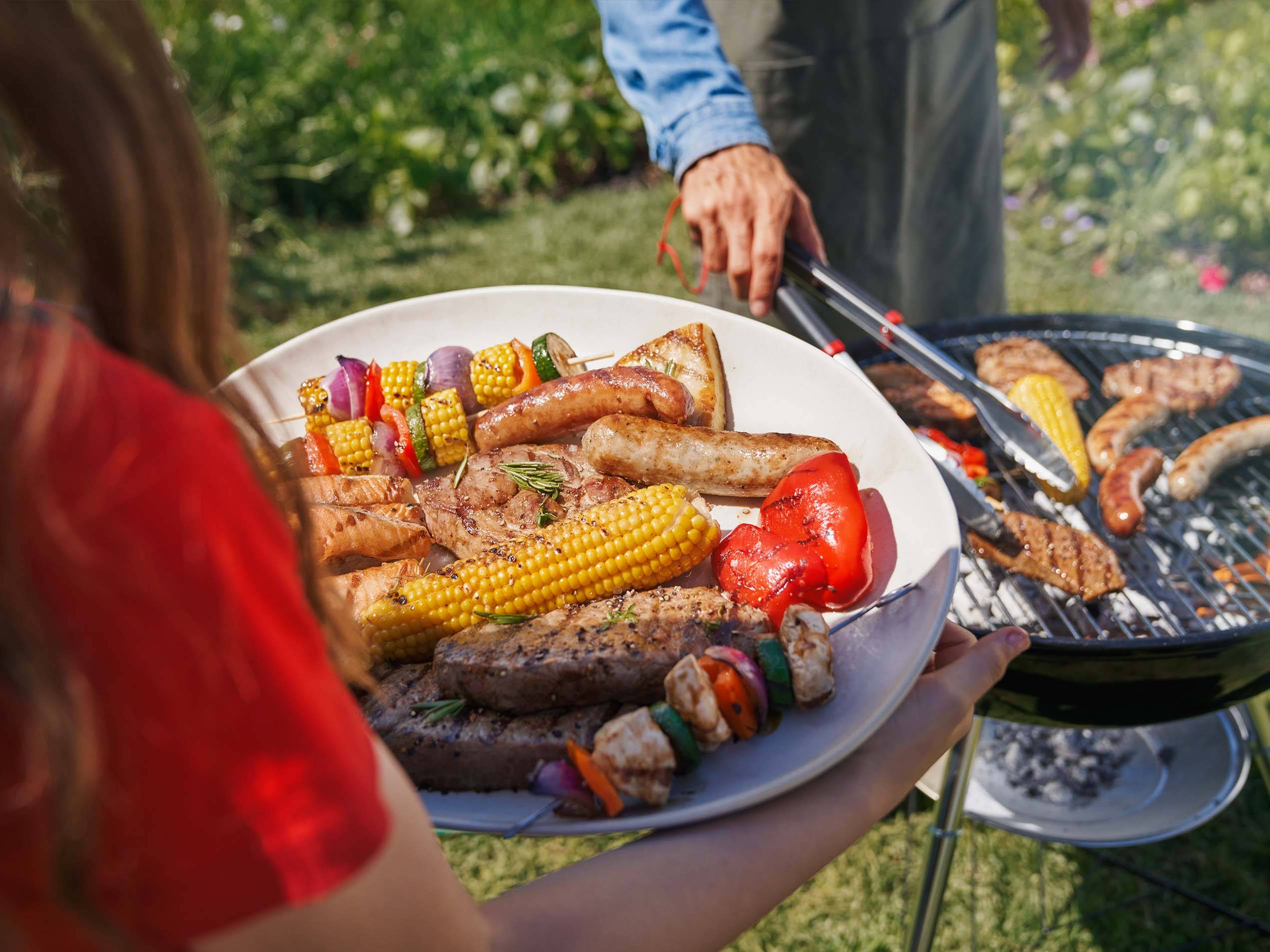 Ein Mädchen mit einem Teller voller Grillfleisch, Würstel und Gemüse im Hintergrund sieht man den Griller